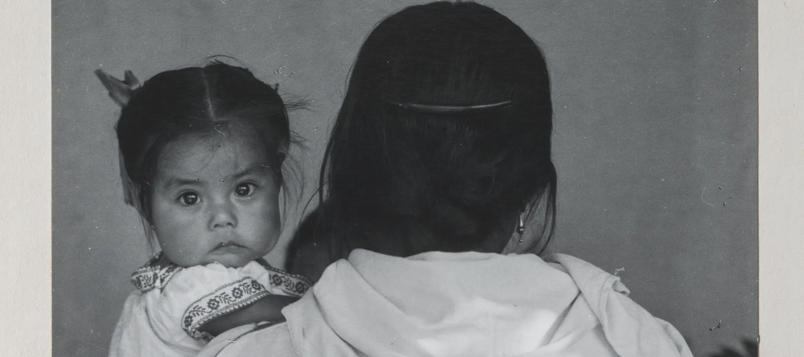 Black-and-white photograph of a woman wearing a dark skirt and light blouse holding a child. The woman faces back and her dark braid reaches past her waist. The dark-haired child stares at the camera and wears a white embroidered dress. Other figures are slightly visible beyond.