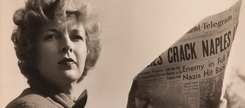 A black-and-white photograph of a light-skinned adult woman holding a newspaper with news about World War II. She wears a coat and her short, curly hair is caught in the wind.