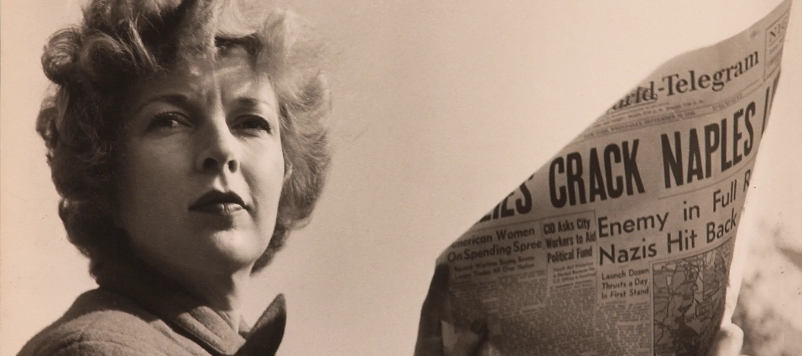 A black-and-white photograph of a light-skinned adult woman holding a newspaper with news about World War II. She wears a coat and her short, curly hair is caught in the wind.