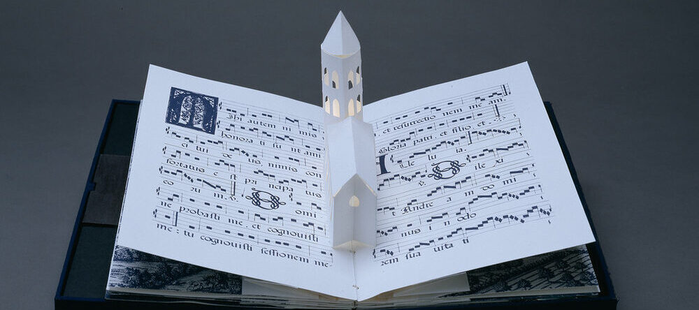 Bound in a dark blue fabric-covered binding, the book opens to display five pop-up towers illuminated from within by a tiny bulb. The featured page includes a white country church with a steeple, set into pages featuring musical scores presented in manuscript style.