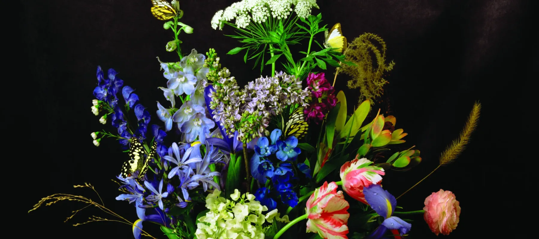 A colorful portrait of a lush arrangement of flowers including poppies, peonies, roses, carnations, and more. Some flowers have small water drpolets on them and a butterfly and bee are perched on others. The image is a still life photograph, though it looks like a painting.