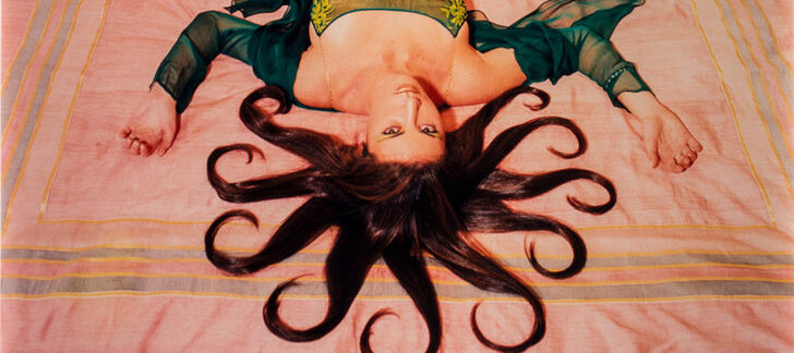 A slim, light-skinned woman wearing a long, emerald-green dress gazes up at the viewer from the foot of the bed, her bare feet near the headboard. Brown hair radiates from her head in 10 curled, snake-like segments. Her arms, bent at the elbows, extend from her body to form a W.
