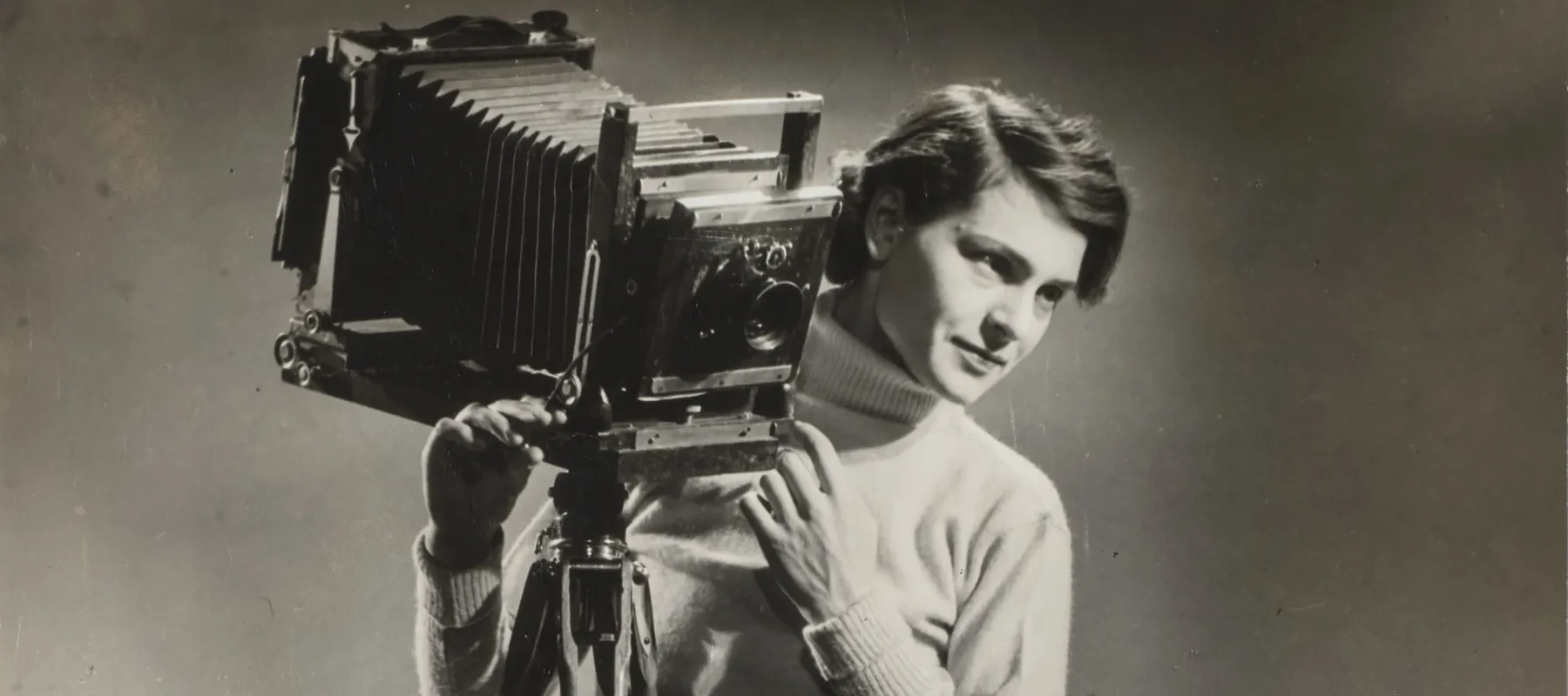 A black-and-white photograph of a light-skinned adult woman standing next to a vintage camera mounted on legs.