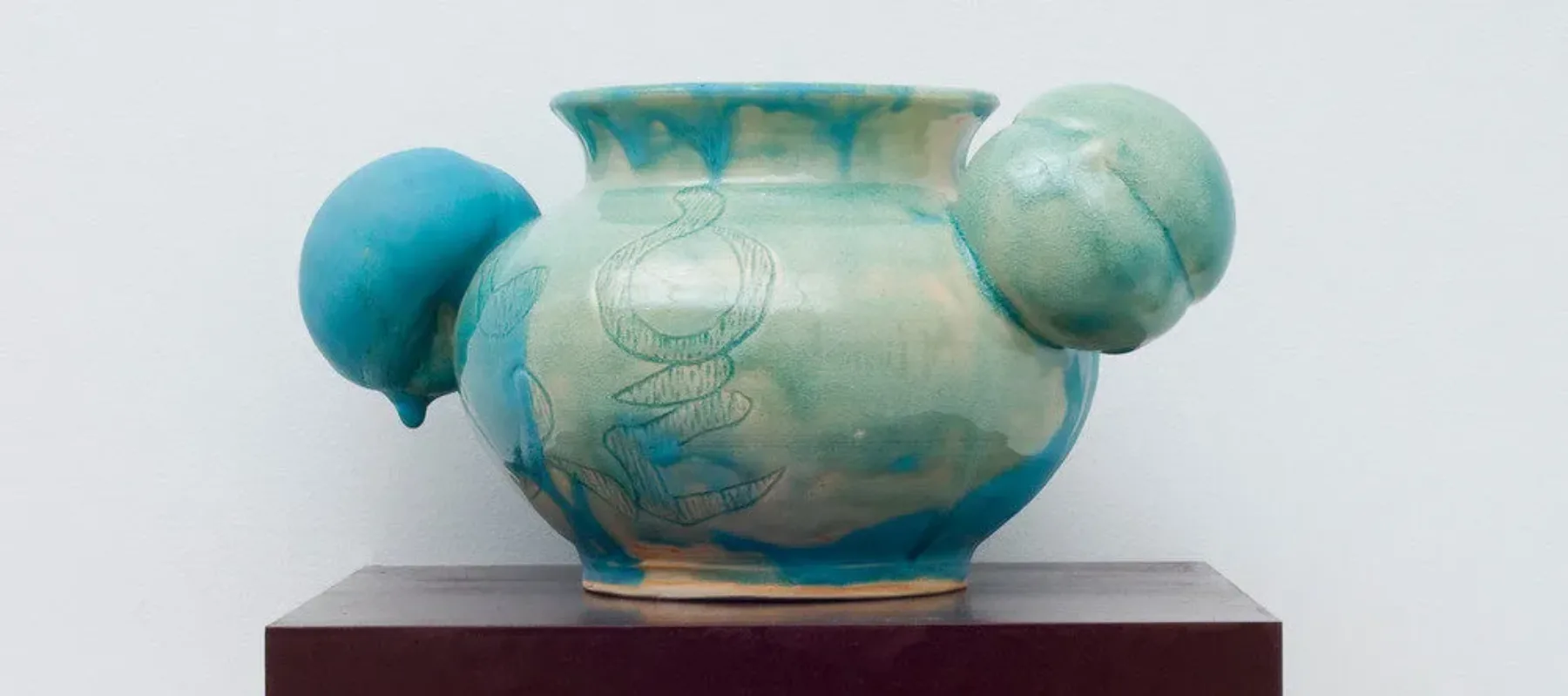 A round, ceramic vessel glazed unevenly in glossy celadon has a bulging body incised with abstract shapes. Matte turquoise glaze dribbling down from the rim obscures portions of the pale green and fully coats one of two peach-shaped orbs projecting from either side of the neck.