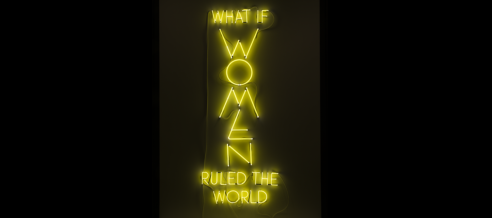 The phrase “What if Women Ruled the World” is sculpted with neon tubes of bright yellow, in all capital letters. The letters of “WOMEN” are larger than the rest and are configured vertically.