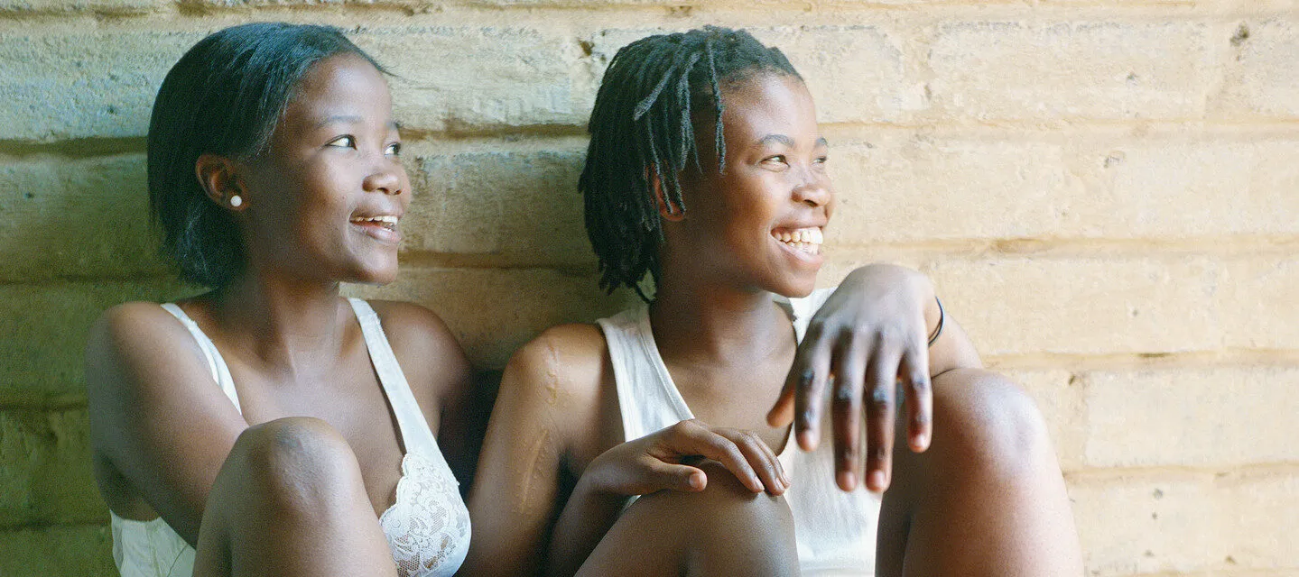 Two dark-skinned young women with short hair sit together against a white brick wall, their limbs lovingly intertwined and glowing smiles across their faces. One wears a white bra and shorts, and the other a white tank-top and blue shorts.