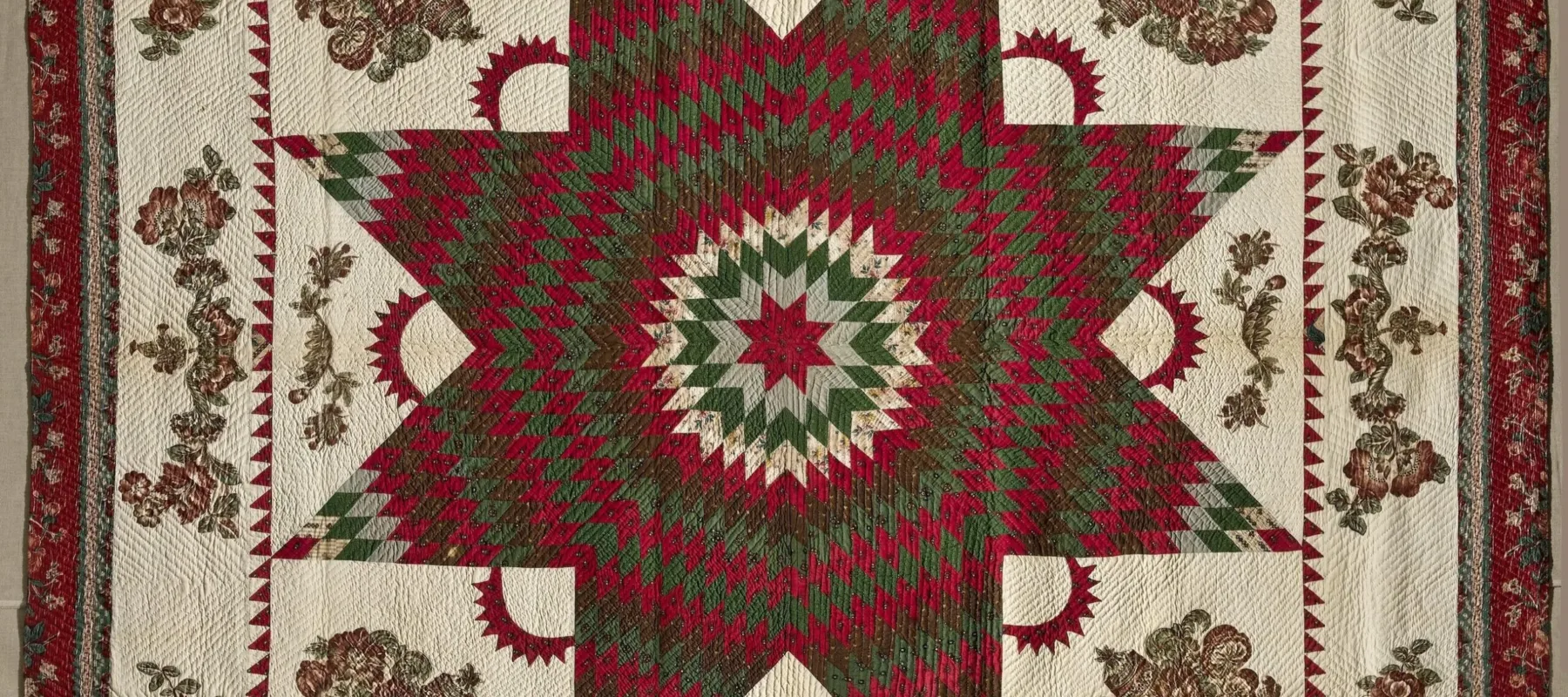 Vintage quilt in dark reds and greens on a cream background. In the center is a large eight-point Star of Bethlehem surrounded by Victorian floral appliques. The border, edged in dark red fabric, features matching floral garland appliques.