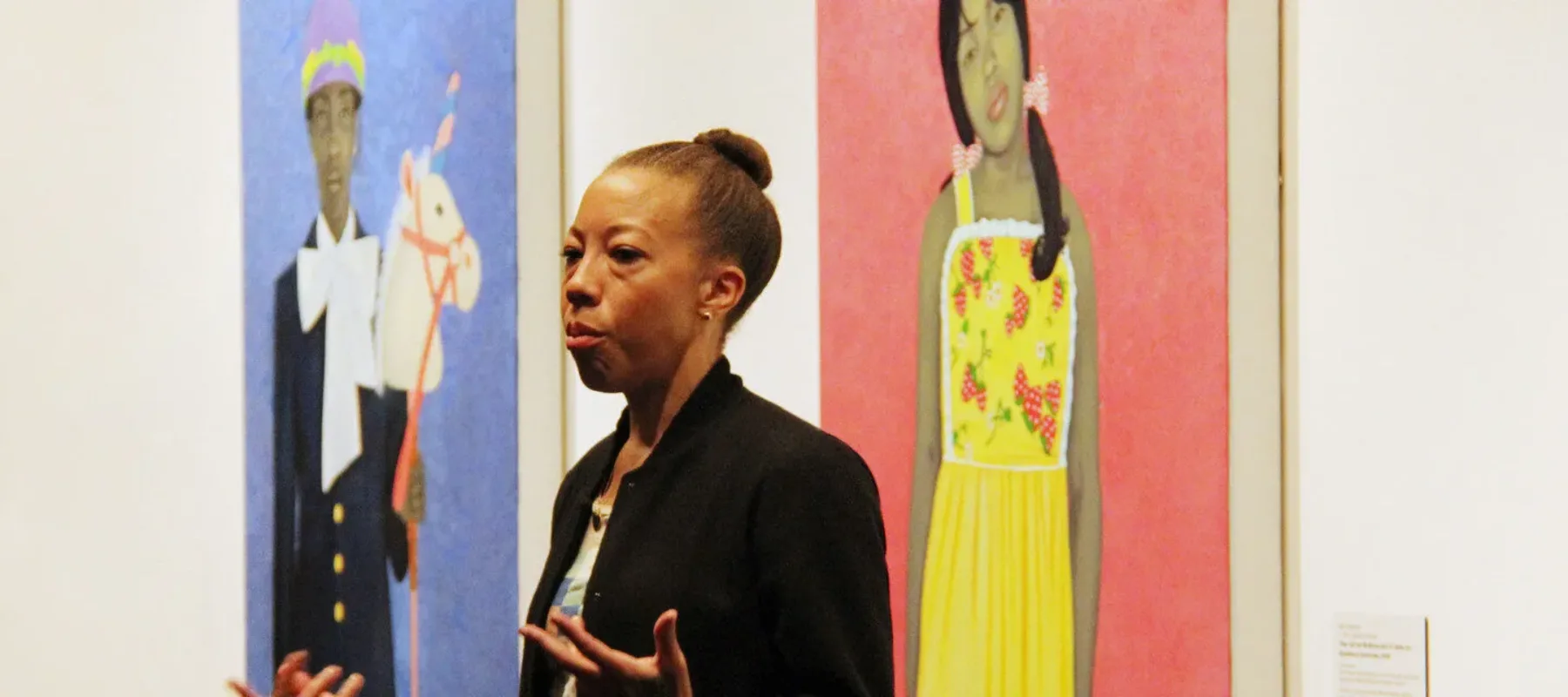 Amy Sherald speaks to a crowd in a gallery beside her striking portraits depicting individuals with gray skintones against vibrant, solid-colored backgrounds. She gestures with her hands and has a serious expression on her face.