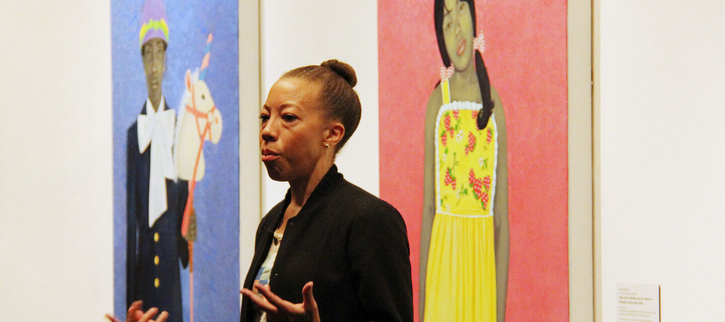 Amy Sherald speaks to a crowd in a gallery beside her striking portraits depicting individuals with gray skintones against vibrant, solid-colored backgrounds. She gestures with her hands and has a serious expression on her face.