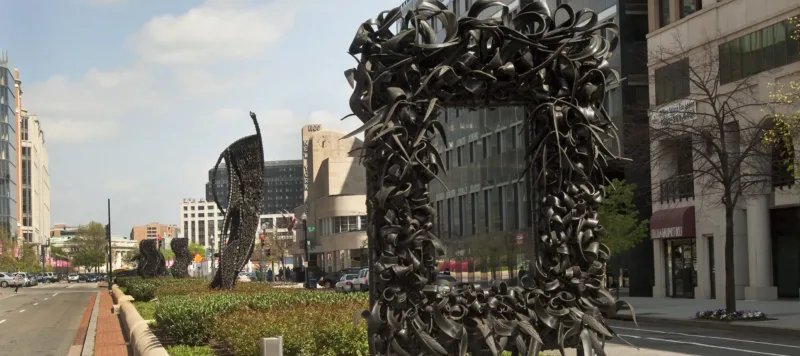 A large metal sculpture stands in the middle of the street, similar to an empty rectangular picture frame. The “frame” is crowded with undulating, coiling metal sculptural elements that occasionally end in spikes. Behind stand three vertical twisting, net-like metal sculptures.