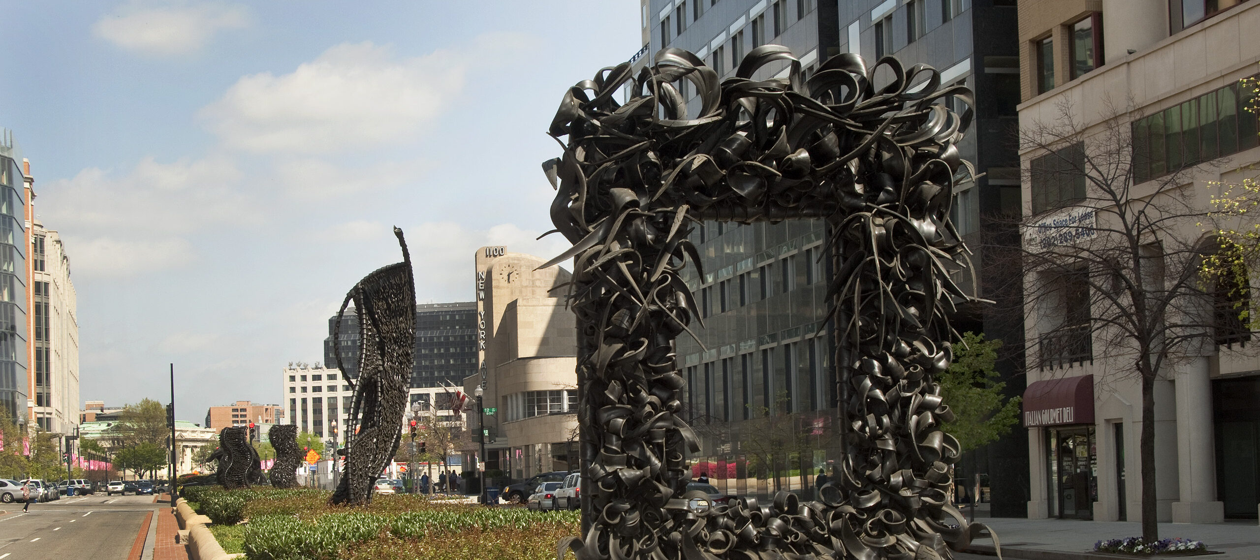 A large metal sculpture stands in the middle of the street, similar to an empty rectangular picture frame. The “frame” is crowded with undulating, coiling metal sculptural elements that occasionally end in spikes. Behind stand three vertical twisting, net-like metal sculptures.
