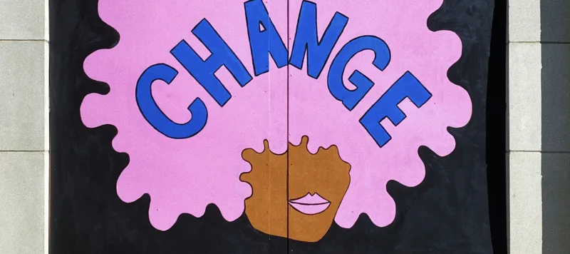 A square plywood mural of a dark-skinned face with a bubblegum pink afro and matching, full lips, against a black background. "CHANGE" is written in blue letters across the afro. The work is signed in the corner “@TRAPXBOB.”