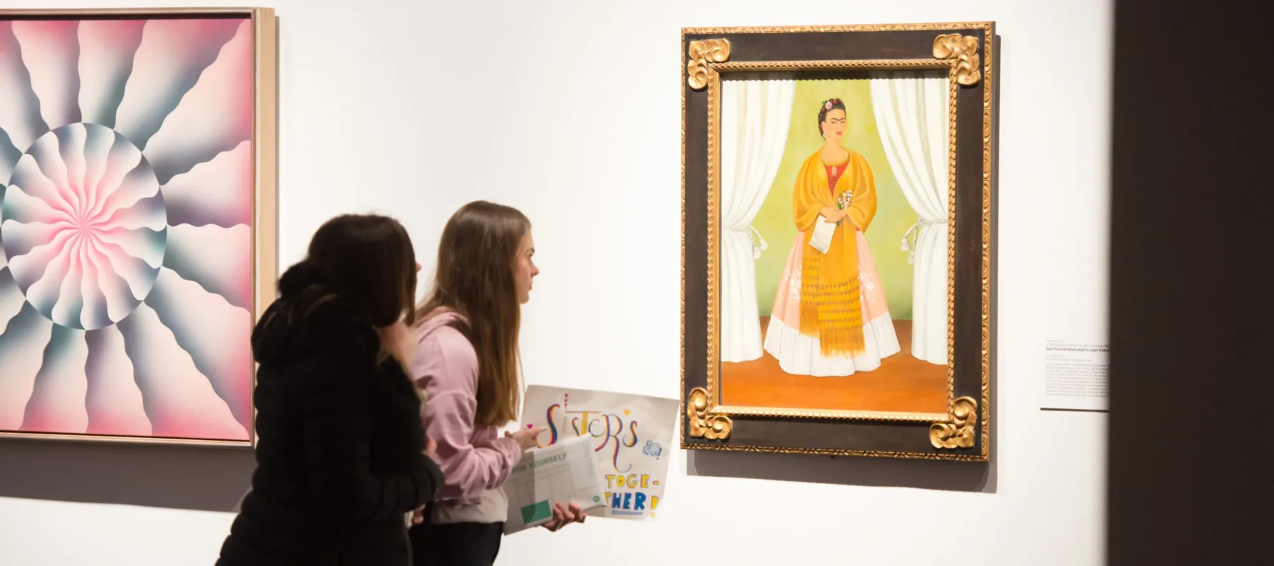 Two young women face away from the camera, craning to get a closer look at a self-portrait by Frida Kahlo