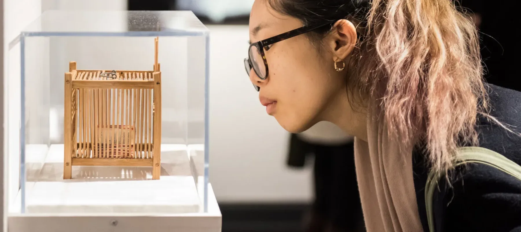 Partial gallery view of a young woman in a ponytail and glasses bending down to look at a tiny contemporary sculpture in a glass vitrine. The sculpture is a wooden cage with an object stored inside. On the back wall hangs a photograph of a light-skinned woman in a white dress.