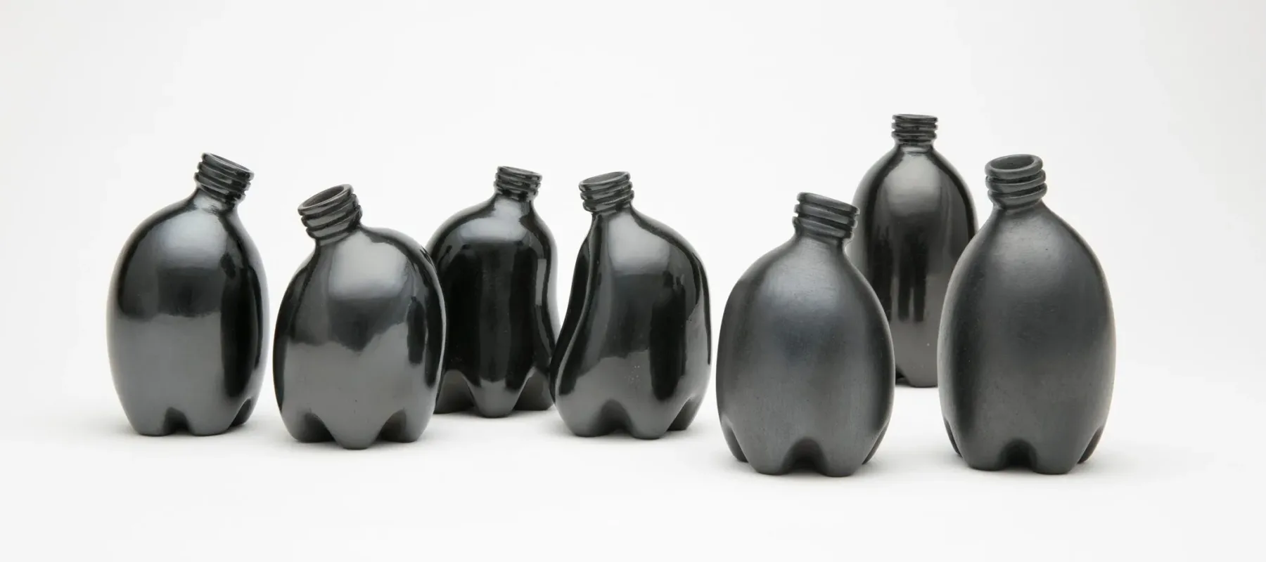 Against a stark white background are seven small clay vessels painted a dark, shiny black. They look like miniature plastic water bottles and are each slightly bent, making them appear like a gathering of people interacting.