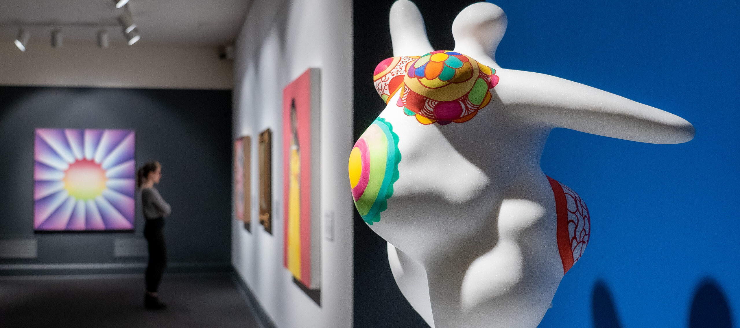 View of the gallery shows an eye-catching marble sculpture in the foreground and a visitor looking at multiple brightly colored artworks in the background. The abstract scupture is of a voluptuous figure with pregnant belly covered in bright patterns and posed with outstretched arms.