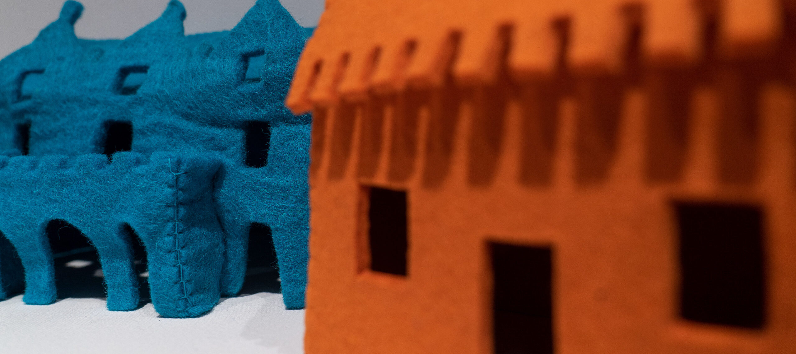 Close-up detail of a larger artwork features a view of two felt houses. On the viewer's right is an orange house that is out-of-focus while on the left is a blue house that is in-focus and has visible stitches, coarse texture, and rectangular windows.
