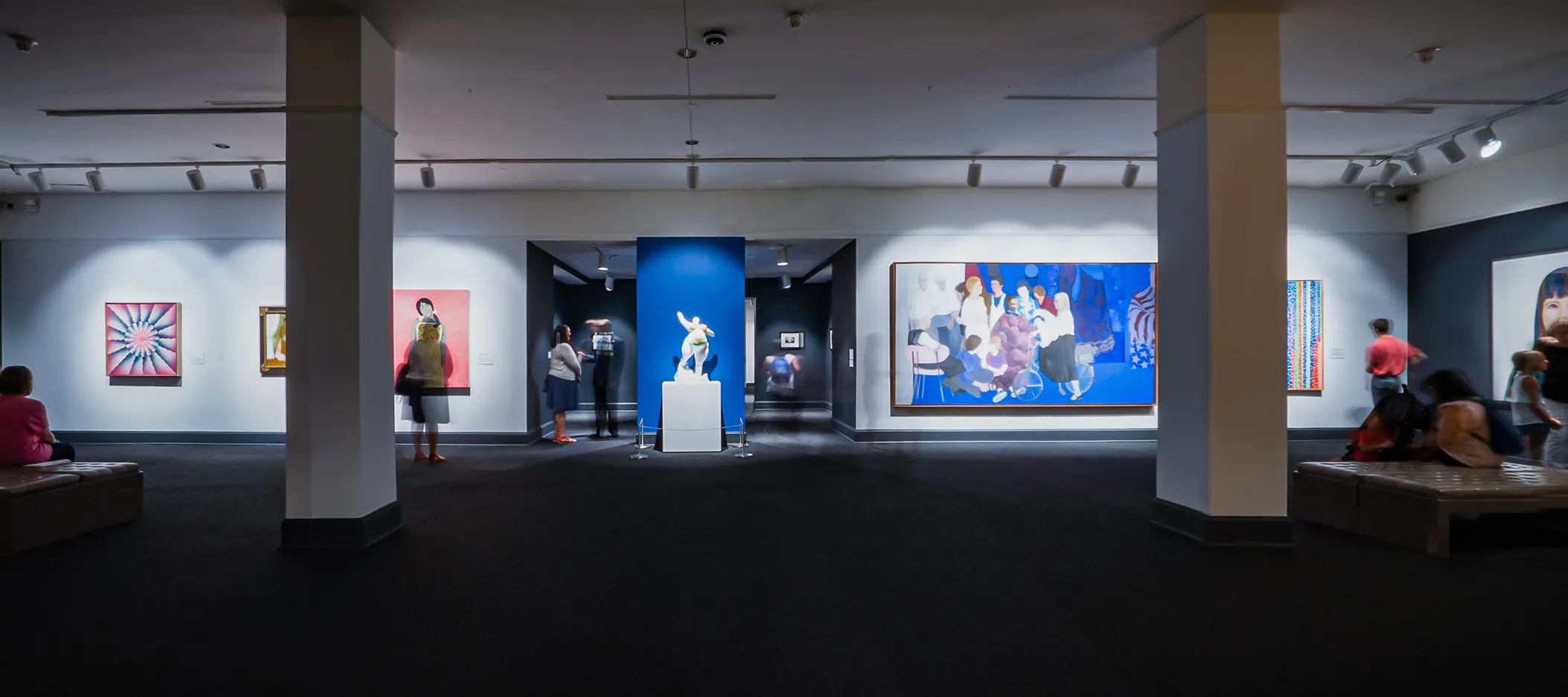 Wide angle view showing a full gallery. The floor is a dark gray carpet and there are two columns, through which large scale works in vibrant colors are visible.