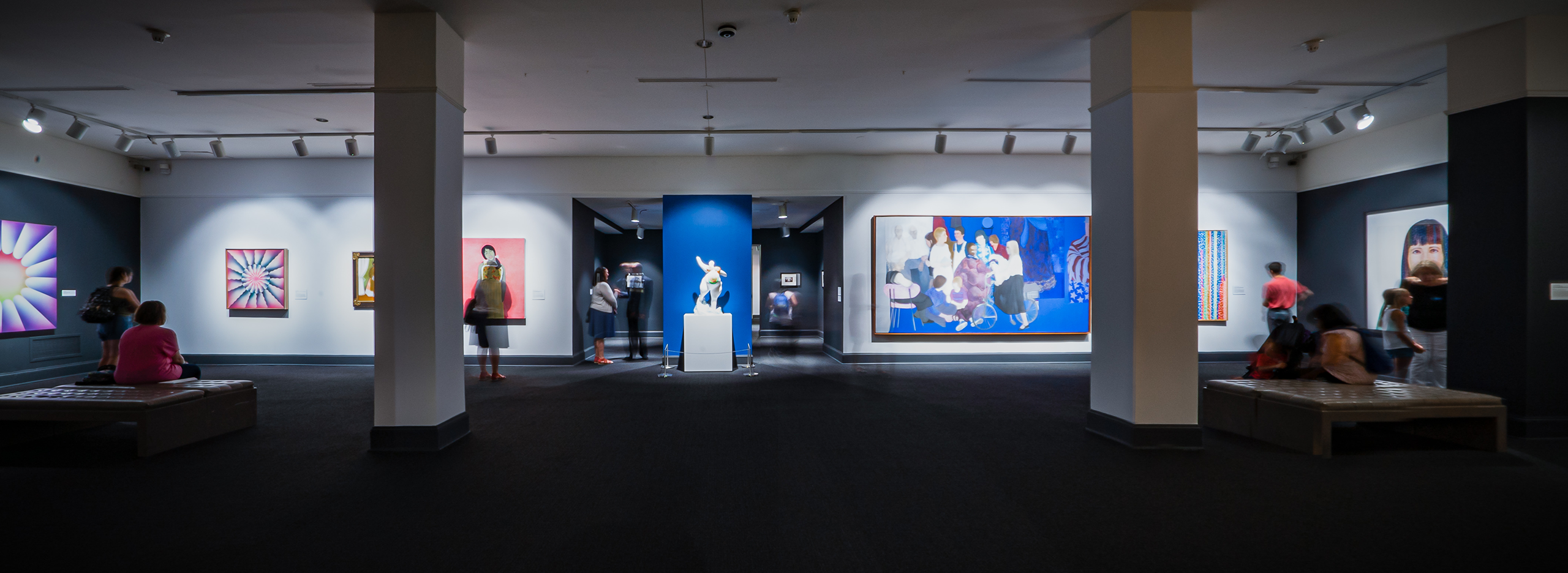 Wide angle view showing a full gallery. The floor is a dark gray carpet and there are two columns, through which large scale works in vibrant colors are visible.