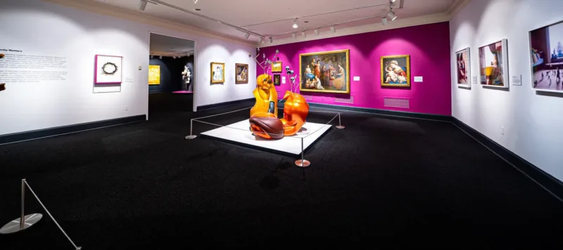 Gallery view with contemporary sculpture made of orange motor scooters that resemble two antlered animals fighting in foreground. Four paintings hang on a magenta wall in background, including a baroque painting of the Virgin and Child, and a portrait of a light-skinned man, women and children gathered together in eighteenth-century attire. To the left on a white wall hang two other paintings.