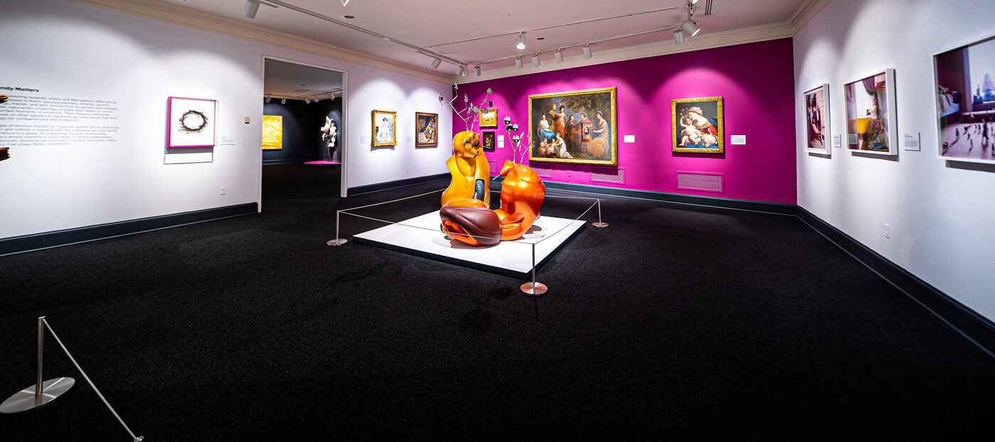 Gallery view with contemporary sculpture made of orange motor scooters that resemble two antlered animals fighting in foreground. Four paintings hang on a magenta wall in background, including a baroque painting of the Virgin and Child, and a portrait of a light-skinned man, women and children gathered together in eighteenth-century attire. To the left on a white wall hang two other paintings.