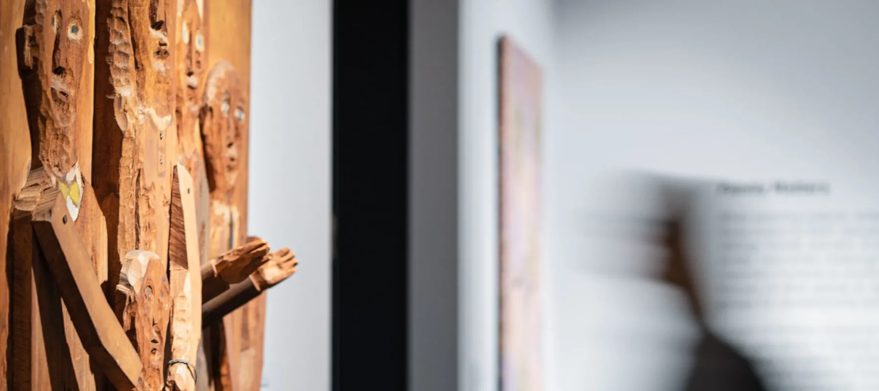 Side view of wooden relief sculpture on wall at left. Relief depicts five figures of various sizes whose arms and hands extend out from sculpture. At right is a blurred figure moving left, heading toward a dark doorway. Behind figure on wall are an artwork and text, out of focus.