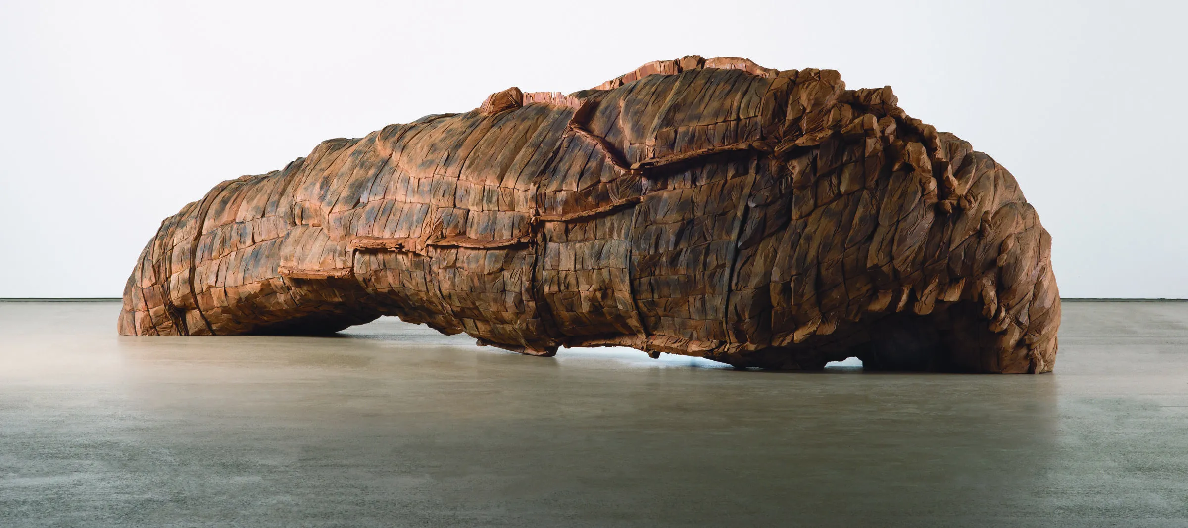 A large cedar and paint sculpture installed against a white wall.