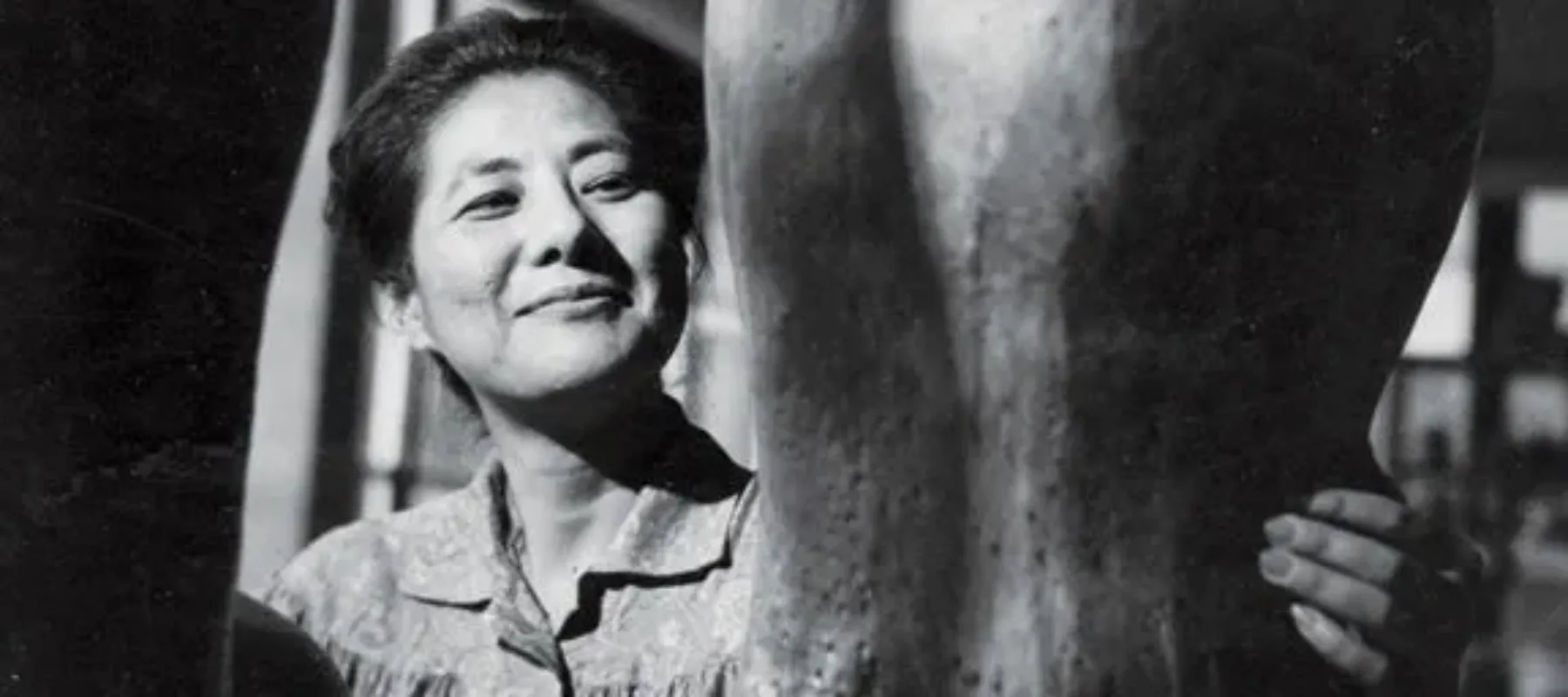 A black-and-white photograph of a light-skinned, dark-haired, Asian adult woman smiling while wearing a smock in her studio. In the foreground are two of her large sculptures; she has her hand on one of them.