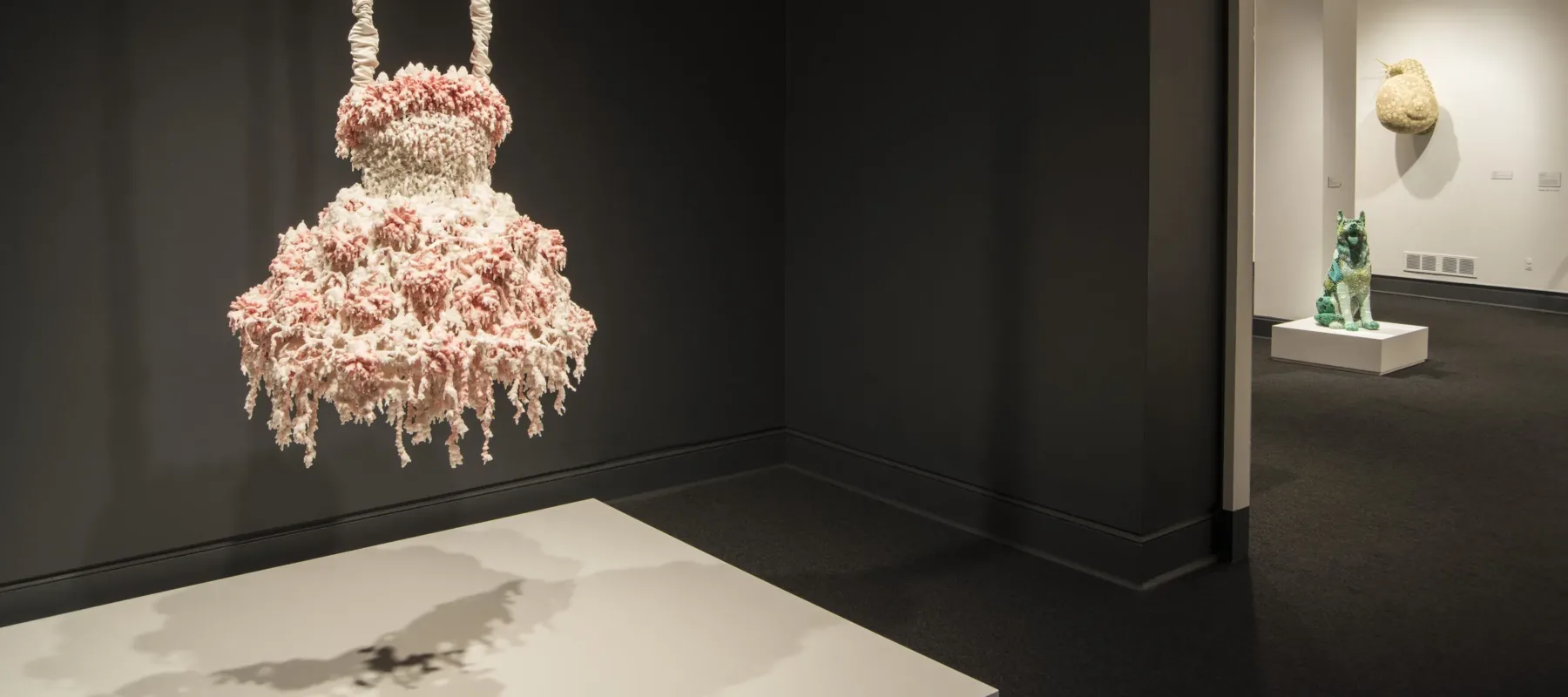 A sculpture hangs in a dark gallery. The sculpture comprises layers of melted pink and white wax that form a dress-like shape hanging from satin-wrapped chains. Its color, shape, and bumpy, lacy texture, evoke a frilly tutu, lavishly frosted wedding cake, or coral.