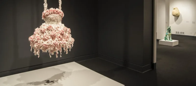 A sculpture hangs in a dark gallery. The sculpture comprises layers of melted pink and white wax that form a dress-like shape hanging from satin-wrapped chains. Its color, shape, and bumpy, lacy texture, evoke a frilly tutu, lavishly frosted wedding cake, or coral.