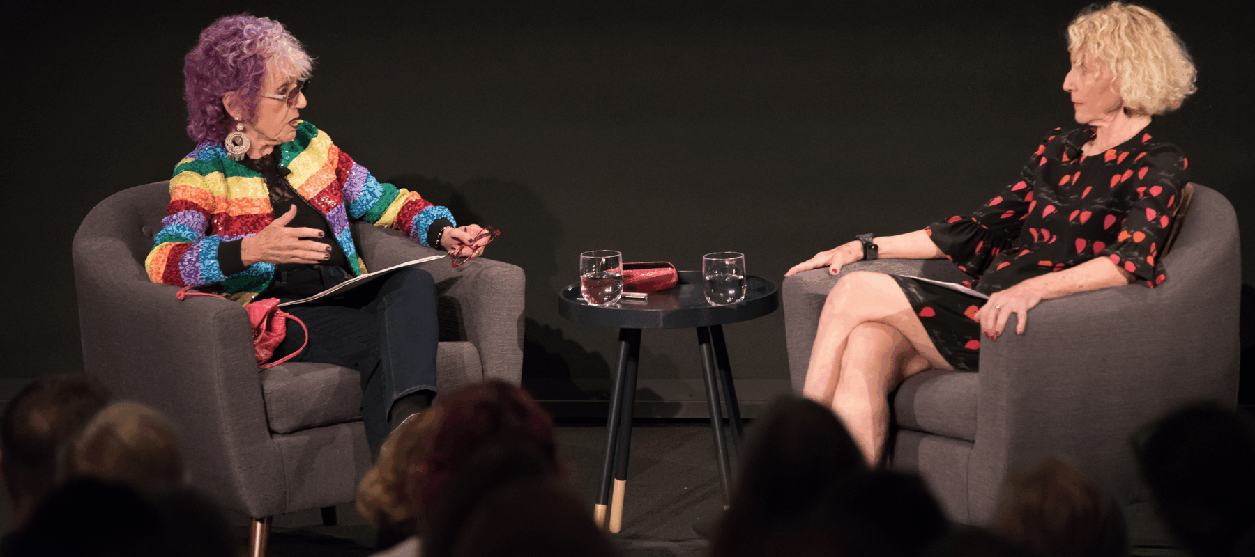 Two women with a light skin tone are sitting across each other on chairs. The woman on the left has purple hair and is wearing a colorful striped jacket.