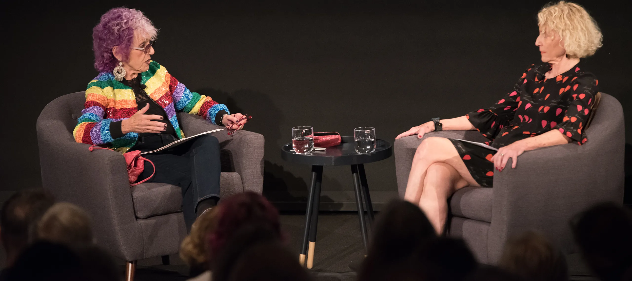 Two women with a light skin tone are sitting across each other on chairs. The woman on the left has purple hair and is wearing a colorful striped jacket.