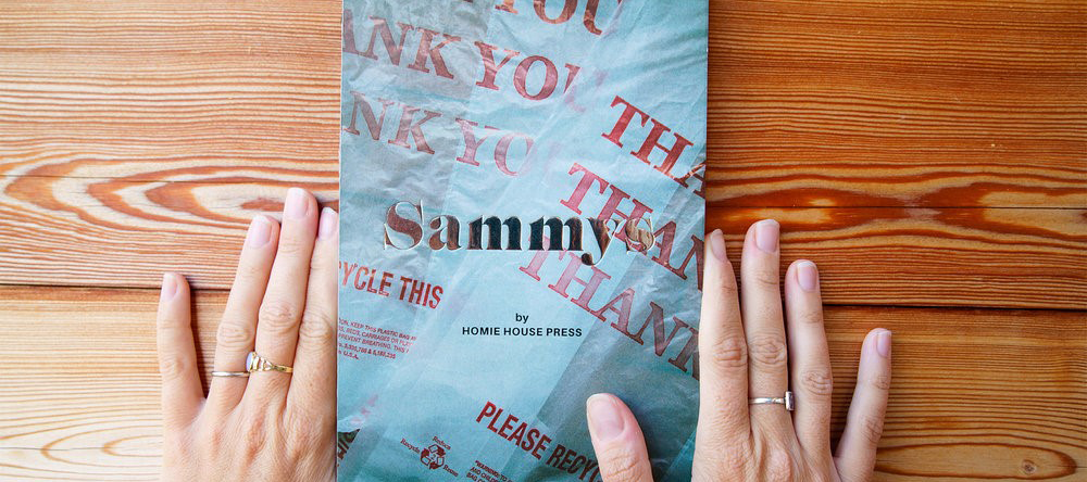 Two hands frame the cover of a book that is titled "Sammys" and the cover is sandwich paper wrapping.