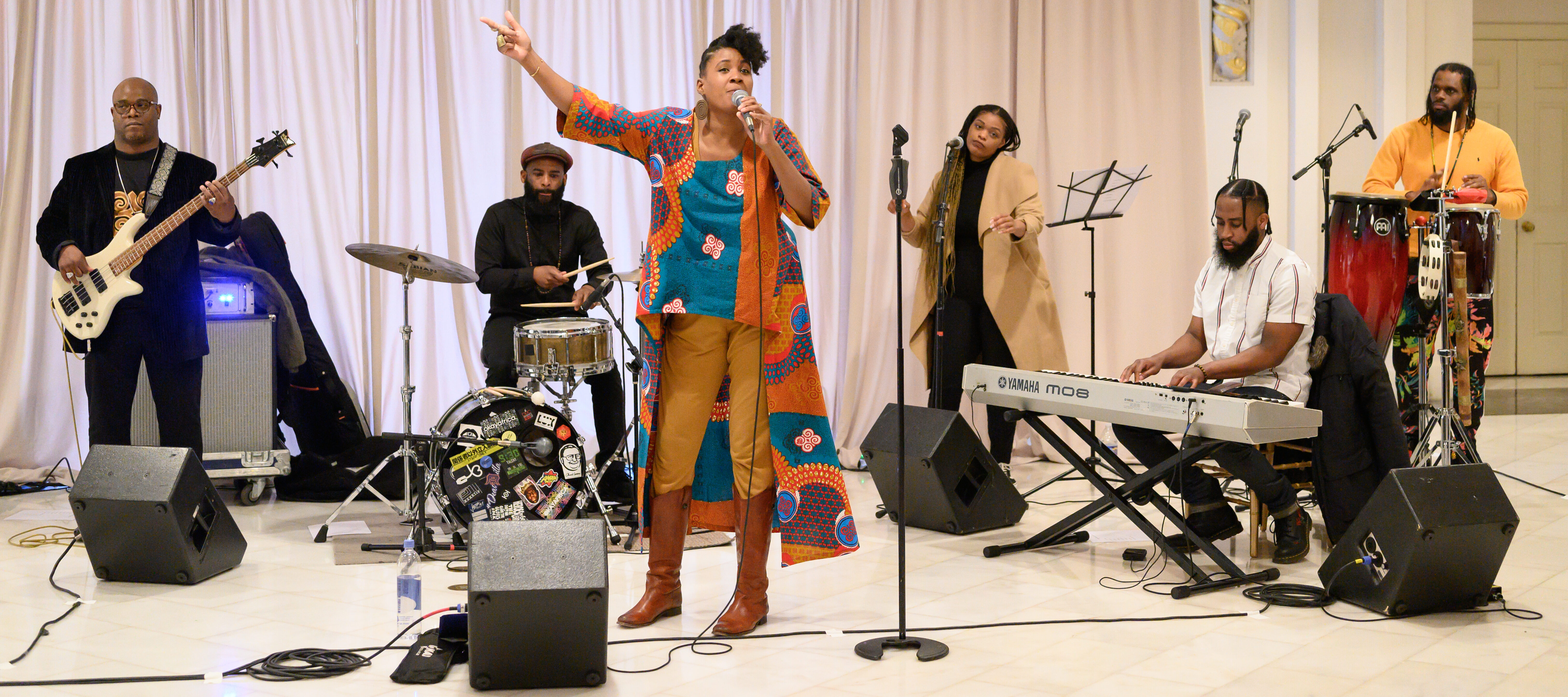 A medium skin-toned adult woman wearing colorful clothing sings into a microphone while a medley of musicians play behind her.