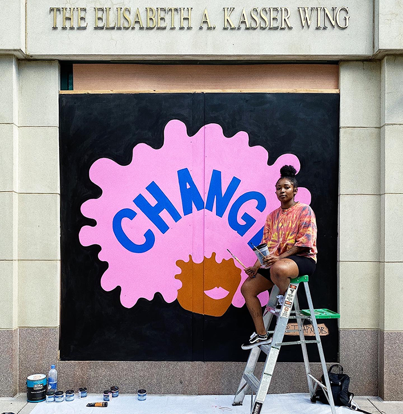 A dark-skinned woman holding paint and paintbrushes sits on a ladder and looks at the camera. Behind her, a mural depicts a dark-skinned stylized head with bright solid pink Afro and lips. The word “change” is painted in navy capital letters across the expanse of hair.