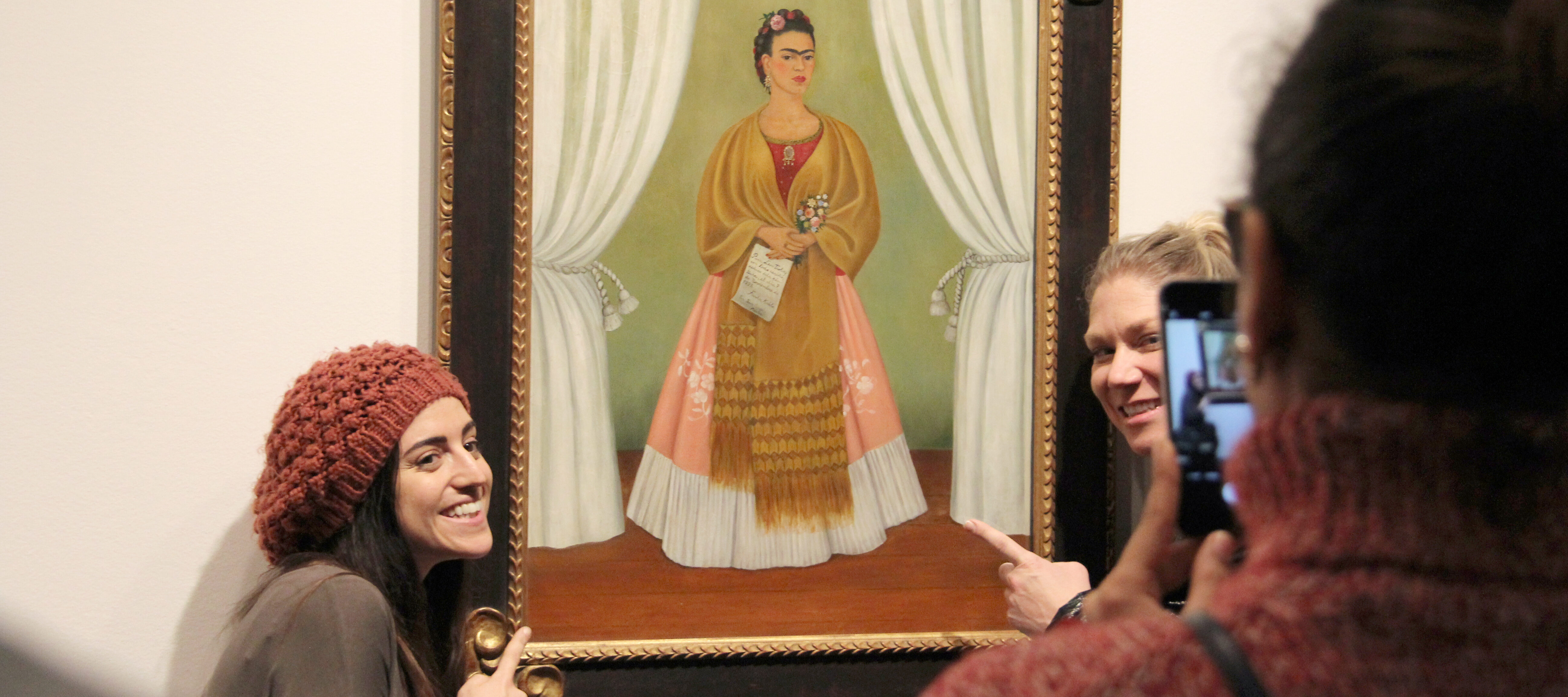 Two light-skinned adults posing for a photo with a painted self-portrait of Frida Kahlo. They are grinning and pointing at the painting, while another person takes their photo on a smartphone.