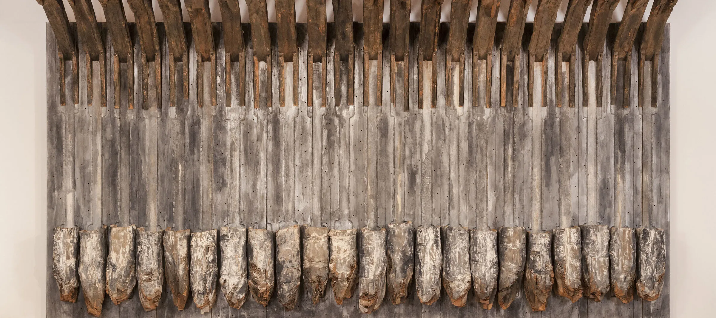 A massive sculpture made of bare cedar slats and painted cedar hunks arranged vertically, flat against a gallery wall.