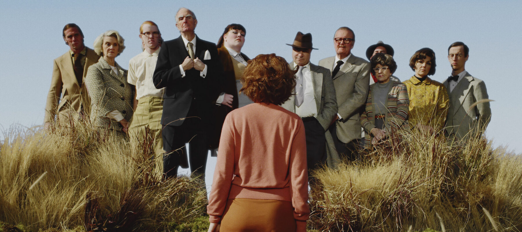 A color photograph of a woman with short red hair and pink cardigan, standing with her back to the camera in a field of tall yellow grass. She faces twelve men and women, who stand slightly elevated above her in a line and look pointedly at her.