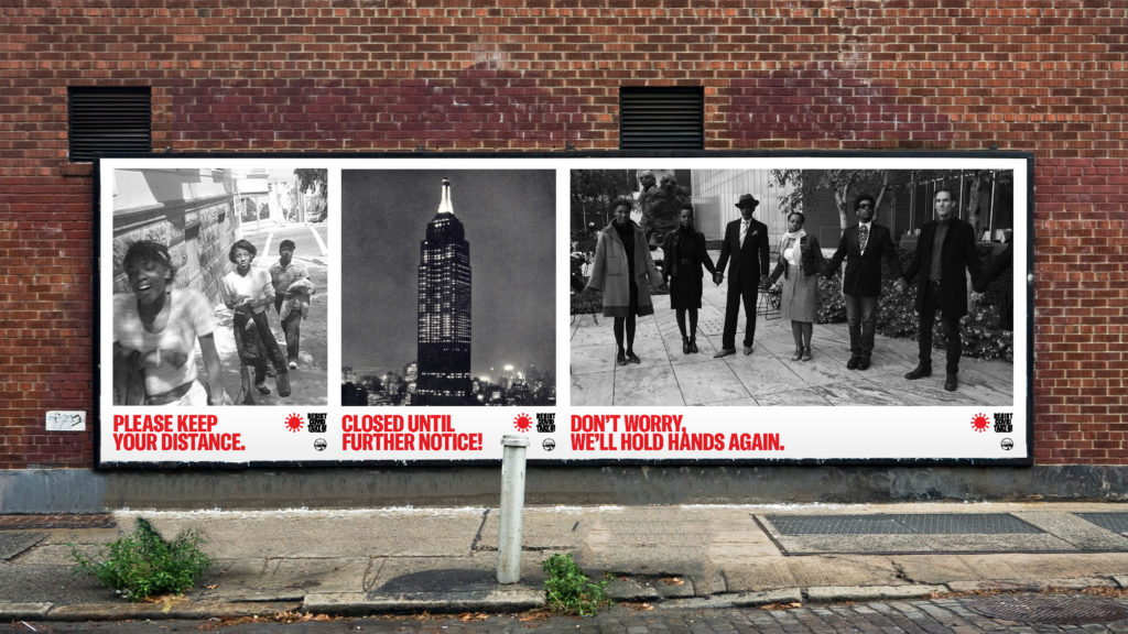 Three posters with black and white photographs occupy a street-level billboard space. The photos are of dark-skinned women standing apart, the Empire State building, and dark-skinned people holding hands. Beneath each is a pandemic-related caption in capital red letters.