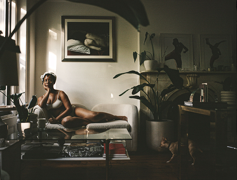 A color photograph of a dark-skinned woman in her underwear, reclining on a love seat. Light streams in from a window on the left, a direction echoed in the diagonal line of the woman’s pose. The rest of the shadowed living room is filled with houseplants, books and art prints. 