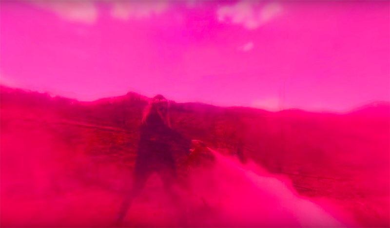 A still from a video, in which a woman with long blond hair and a flower crown stands in front of a mountain range. She assumes a powerful stance as she sprays what looks to be a fire extinguisher. The image has a deep pink haze, as though taken from behind a pink filter.