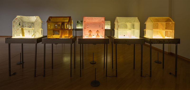 Five small models of houses, each a different muted pastel color, are displayed a dimly lit gallery room on separate hip-height tables. They are lit from within, and each is somewhat translucent; the resulting effect is that they seem to glow from inside.