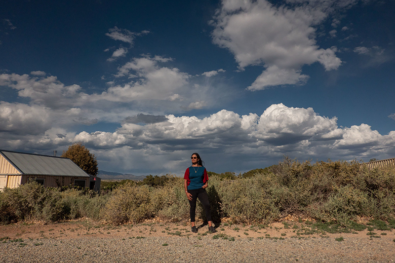 A woman stands on the side of a gravel road. She faces the camera with one hand in her pocket and looks into the distance as the wind blows her long black hair. A house stands in a field of dense shrubbery behind her; a wide blue sky with dramatic cloud stretches above her.