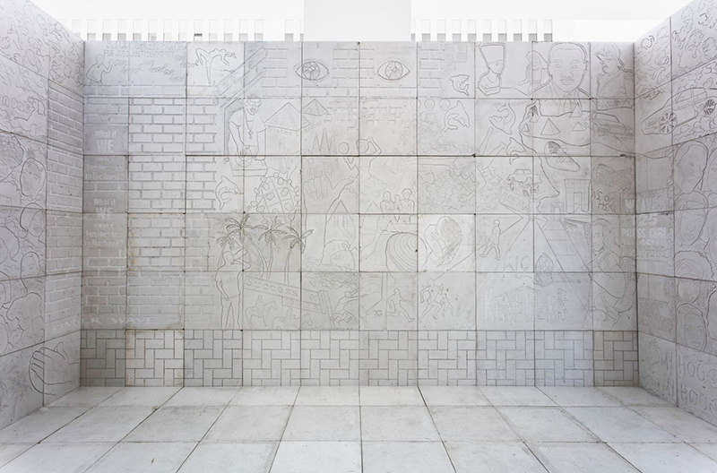 Square blocks of white marble create an open-ceilinged room in the middle of a gallery. The marble walls are inscribed with outlines of overlapping figures, objects, and animals, including Egyptian pyramids, Martin Luther King, Jr. and the bust of Nefertiti.