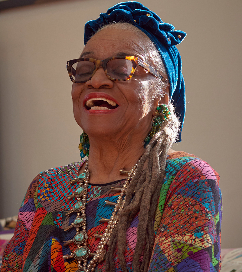 An older dark-skinned woman laughs with her eyes closed and head tilted back. She wears a colorful patterned tunic, a chunky beaded necklace, dangling blue and green earrings, thick glasses, and a blue headscarf that is knotted at the crown of her head.