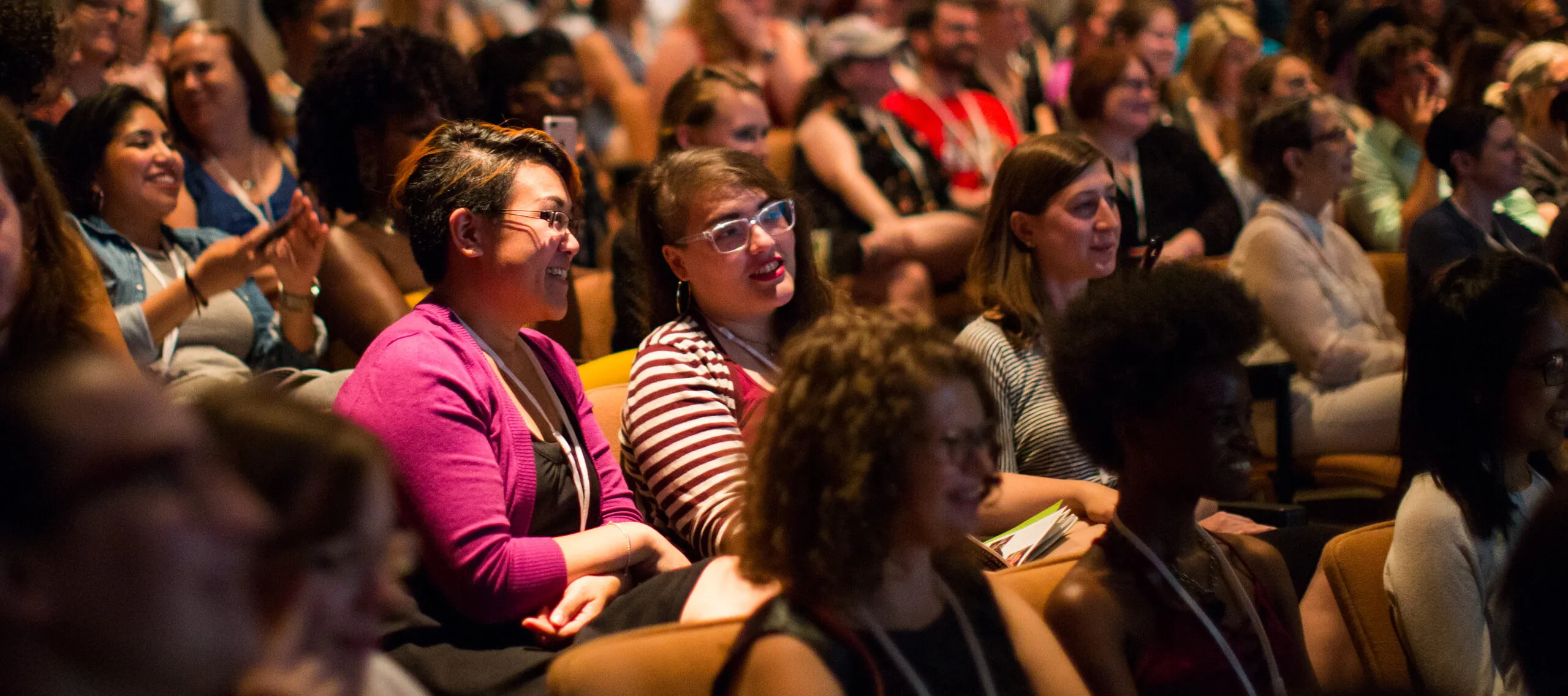 A color photograph of a brightly lit auditorium. Smiling women take up almost every seat. Some are taking photos of the stage with their phone; others lean together in conversation.