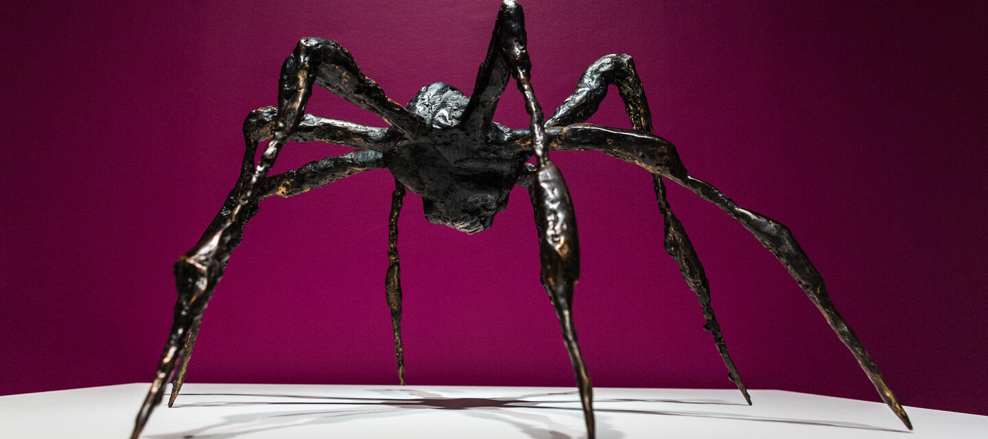 Bronze sculpture of a spider on a white platform against a magenta background.