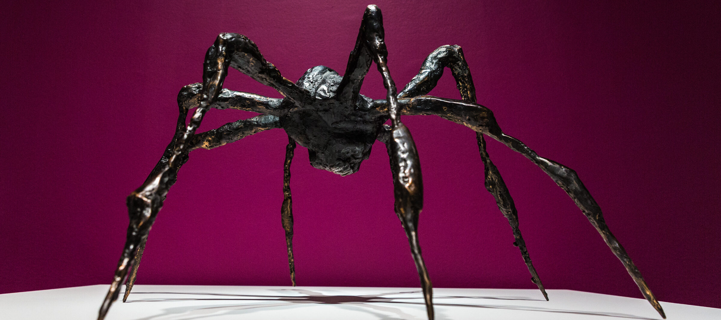 Bronze sculpture of a spider on a white platform against a magenta background.