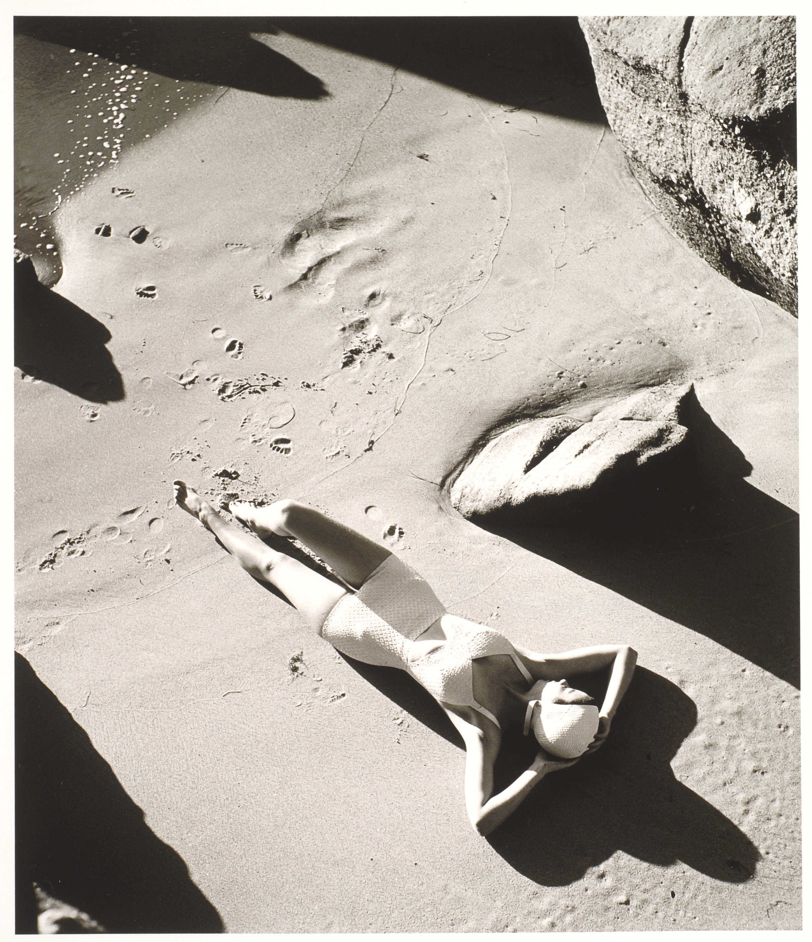 A black and white photograph of a light-skinned woman wearing a one-piece white bathing suit and white swim cap. She is lying down on sand with her hands behind her head. There are footprints in the sand below her, and the image is framed by large rocks.