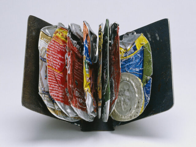 A collection of crushed and flattened aluminum beverage cans displayed in a manner resembling the pages of an open book. The cans are various colors, including red, yellow, and silver, and are arranged vertically between two black, book-like ends.
